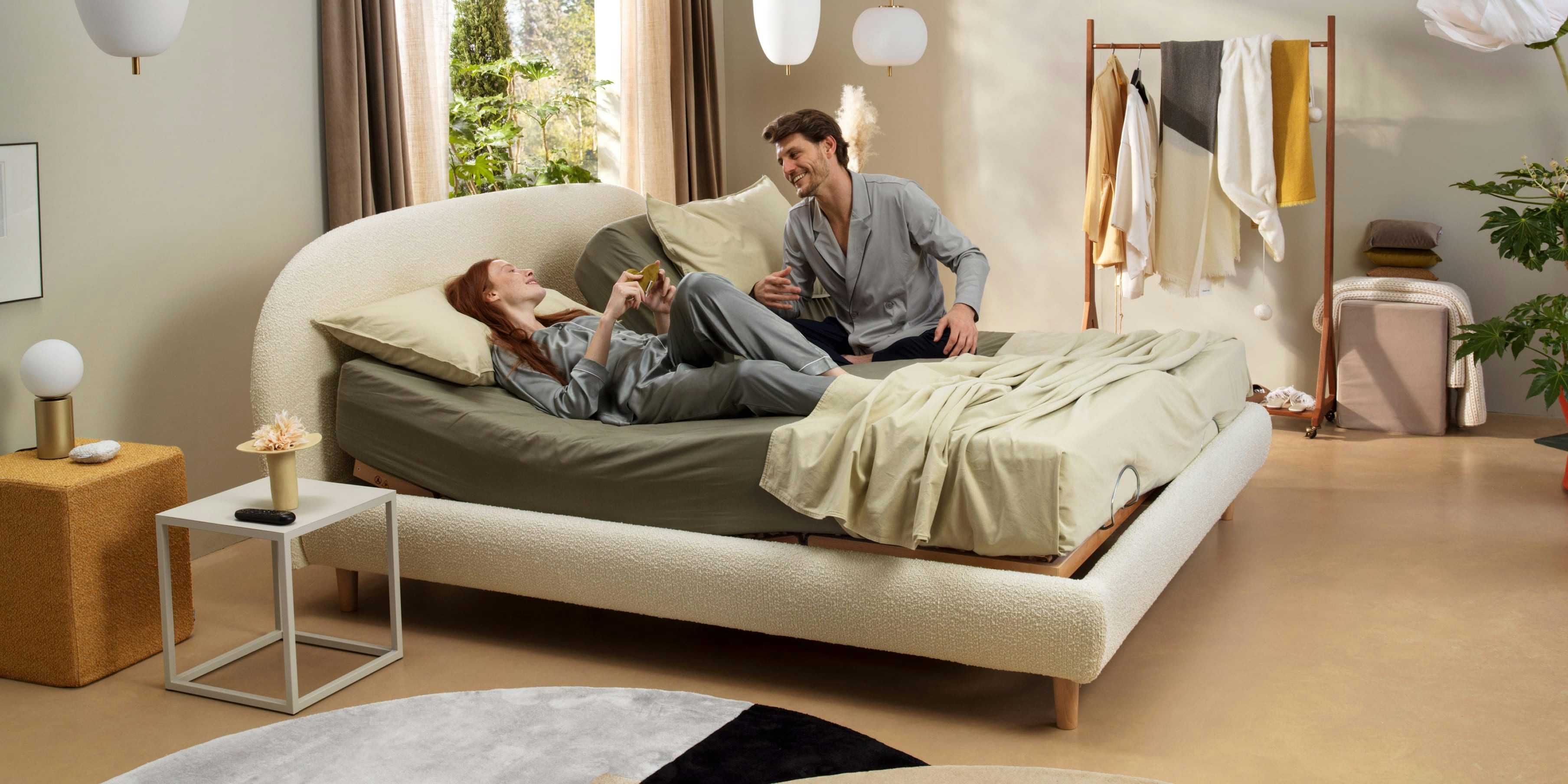 Coppia con pigiami grigi su letto doppio beige in stanza luminosa con pavimento chiaro e arredamento moderno.