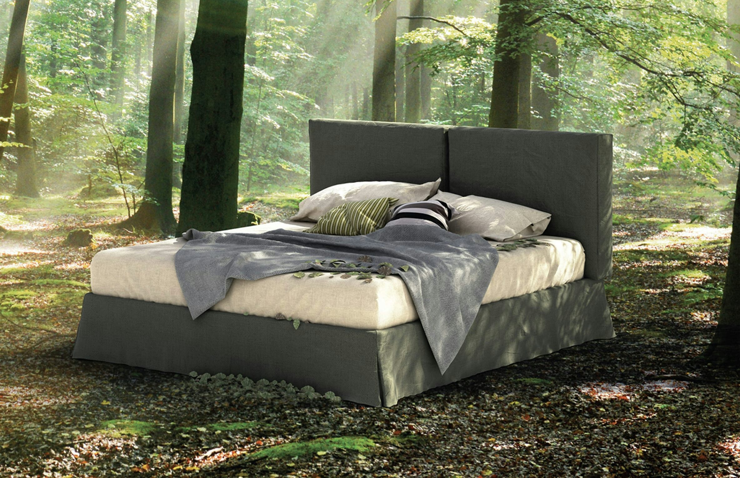 Letto matrimoniale con coperta e cuscini grigi in una foresta illuminata da raggi di sole mattutini.