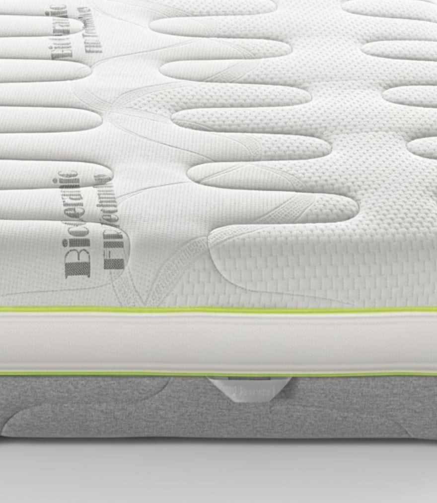 Dettaglio di materasso bianco con trama e scritta bioderica firmata, bordo con cucitura verde lime e base grigia.