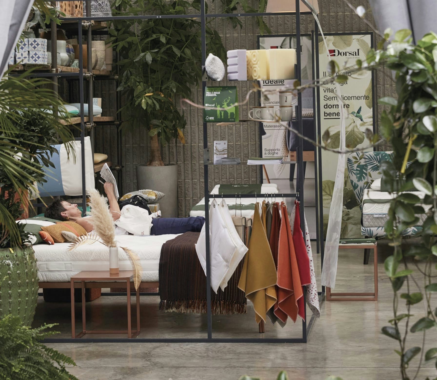 Persona distesa su un letto in esposizione con tessuti colorati appesi e piante verdi intorno in negozio.