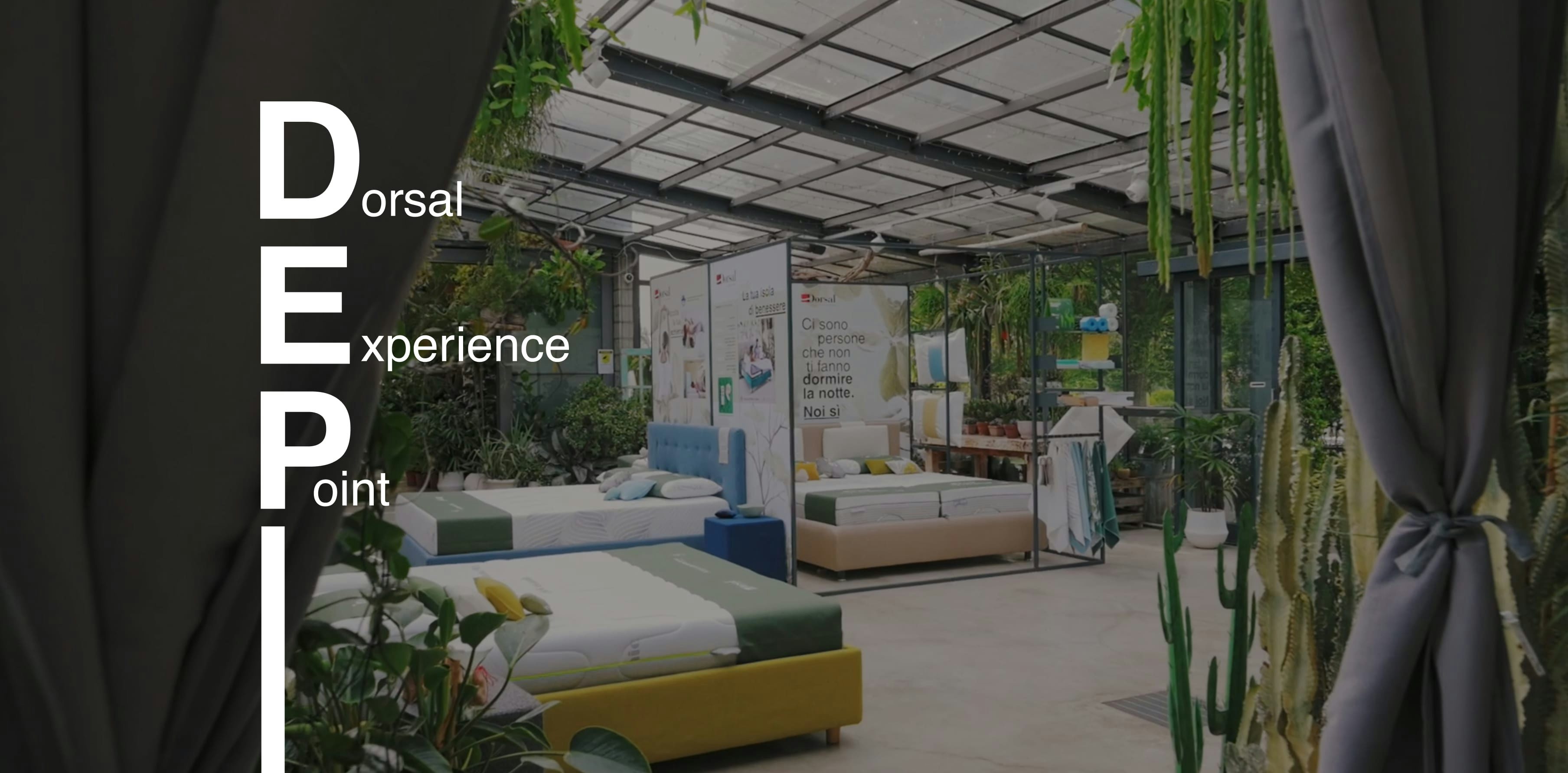 Showroom con letti moderni e piante, ambiente luminoso con grandi finestre e soffitto in vetro, testo Dorsal Experience…