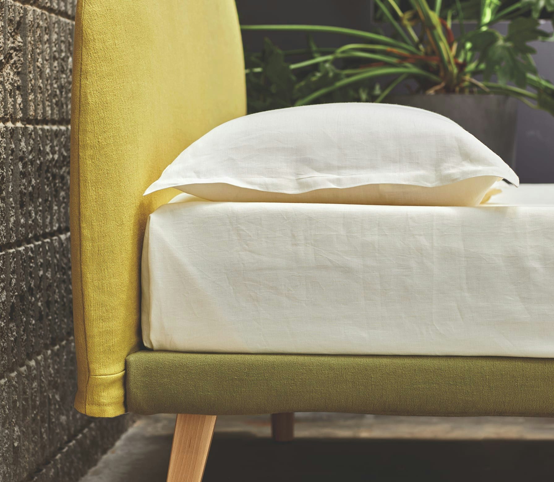 Letto con testiera gialla, base verde e lenzuola bianche con cuscino, accanto parete in mattoni grigi e pianta verde.