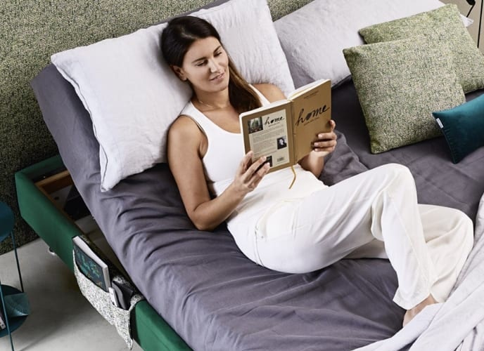 Donna sdraiata su letto verde con lenzuola grigie che legge un libro dal titolo home in ambiente moderno e luminoso.