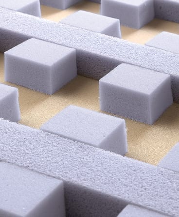 Blocchi e strisce di materiale spugnoso grigio disposti su base beige, texture morbida e superficie uniforme.