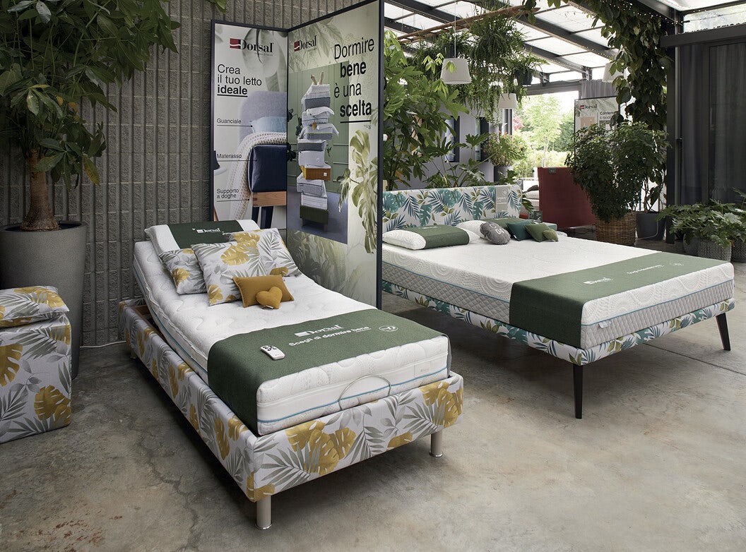 Due letti con materasso e rivestimento a foglie verdi e gialle in showroom con piante e parete divisoria con scritte Dormire…