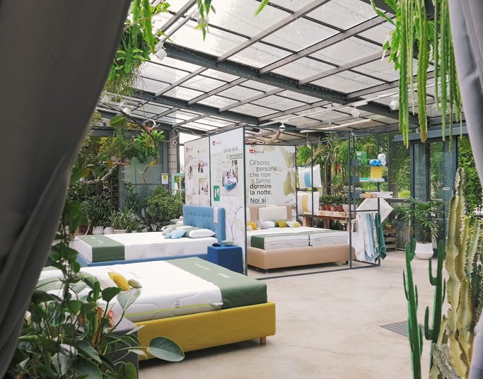 Negozio con letti esposti in ambiente luminoso pieno di piante e pareti di vetro, tende grigie aperte ai lati.