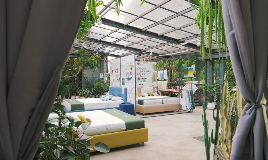 Negozio con letti esposti in ambiente luminoso pieno di piante e pareti di vetro, tende grigie aperte ai lati.
