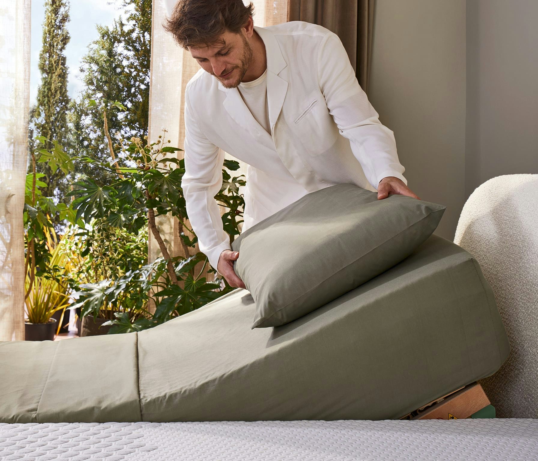 Uomo che sistema un cuscino verde su un letto regolabile con testiera imbottita e piante verdi sullo sfondo.