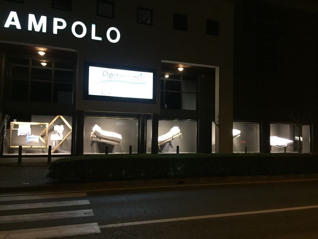 Vetrine illuminate di negozio con letti ortopedici inclinati esposti di notte lungo strada con passaggio pedonale.