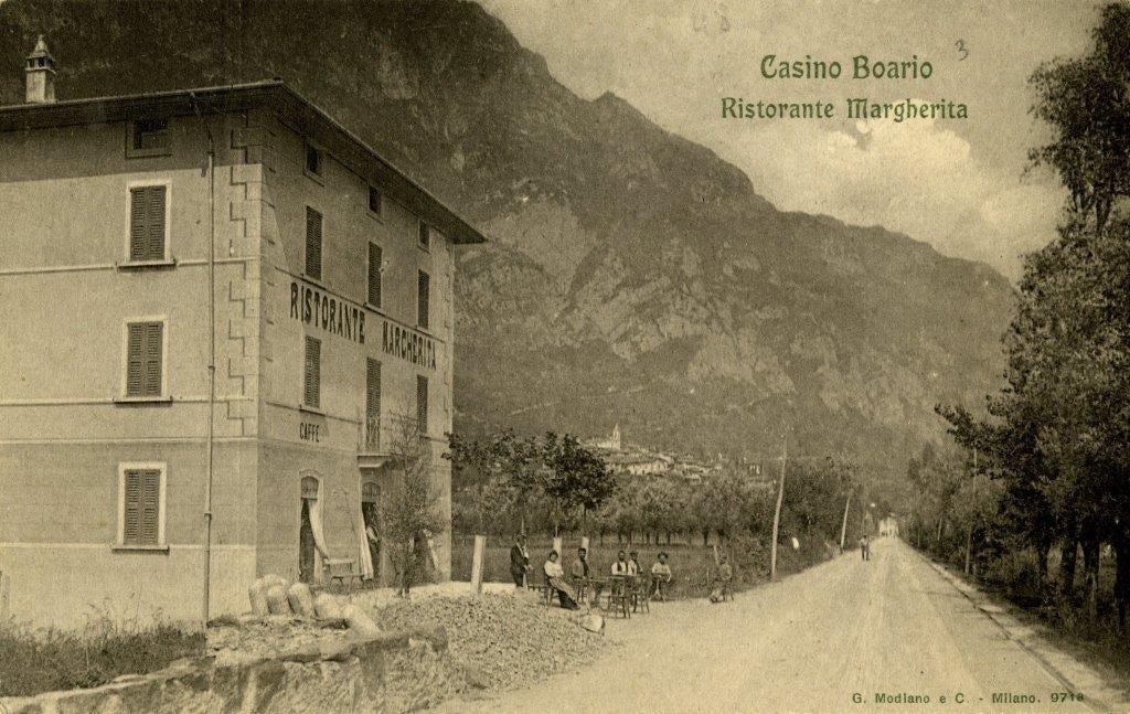 Vecchio ristorante Margherita accanto a strada sterrata con persone sedute e montagna sullo sfondo.