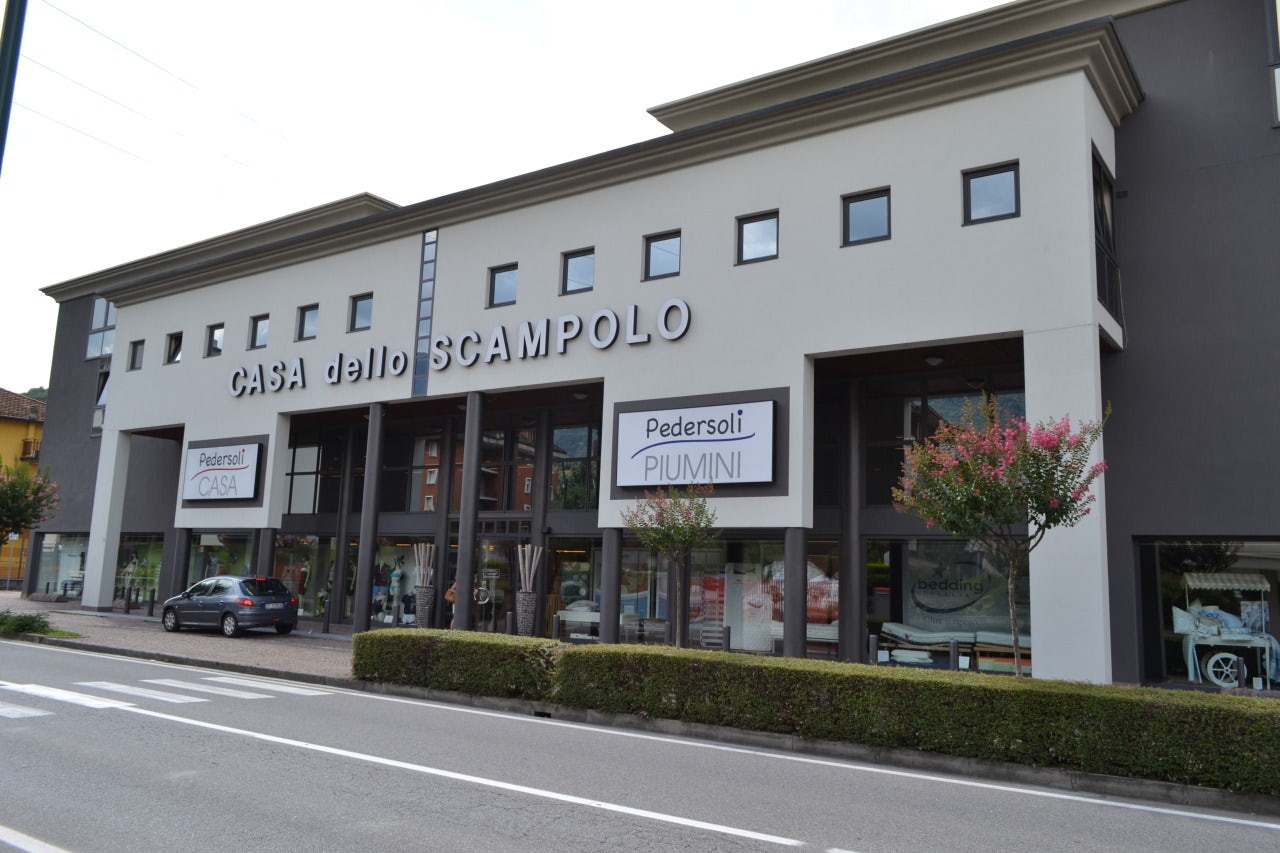Edificio commerciale bianco con insegne Casa dello Scampolo e Pedersoli Piumini, auto parcheggiata e alberi fioriti davanti.