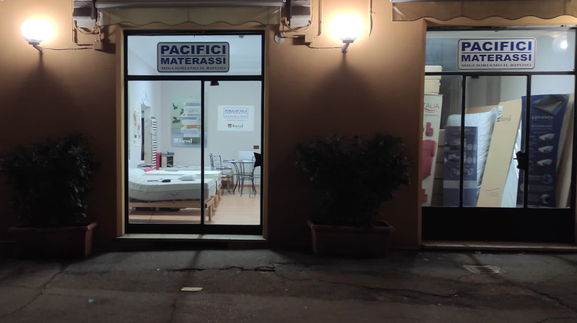 Vetrina notturna del negozio Pacifici Materassi con letti e materassi visibili all'interno e due lampade accese all'esterno.