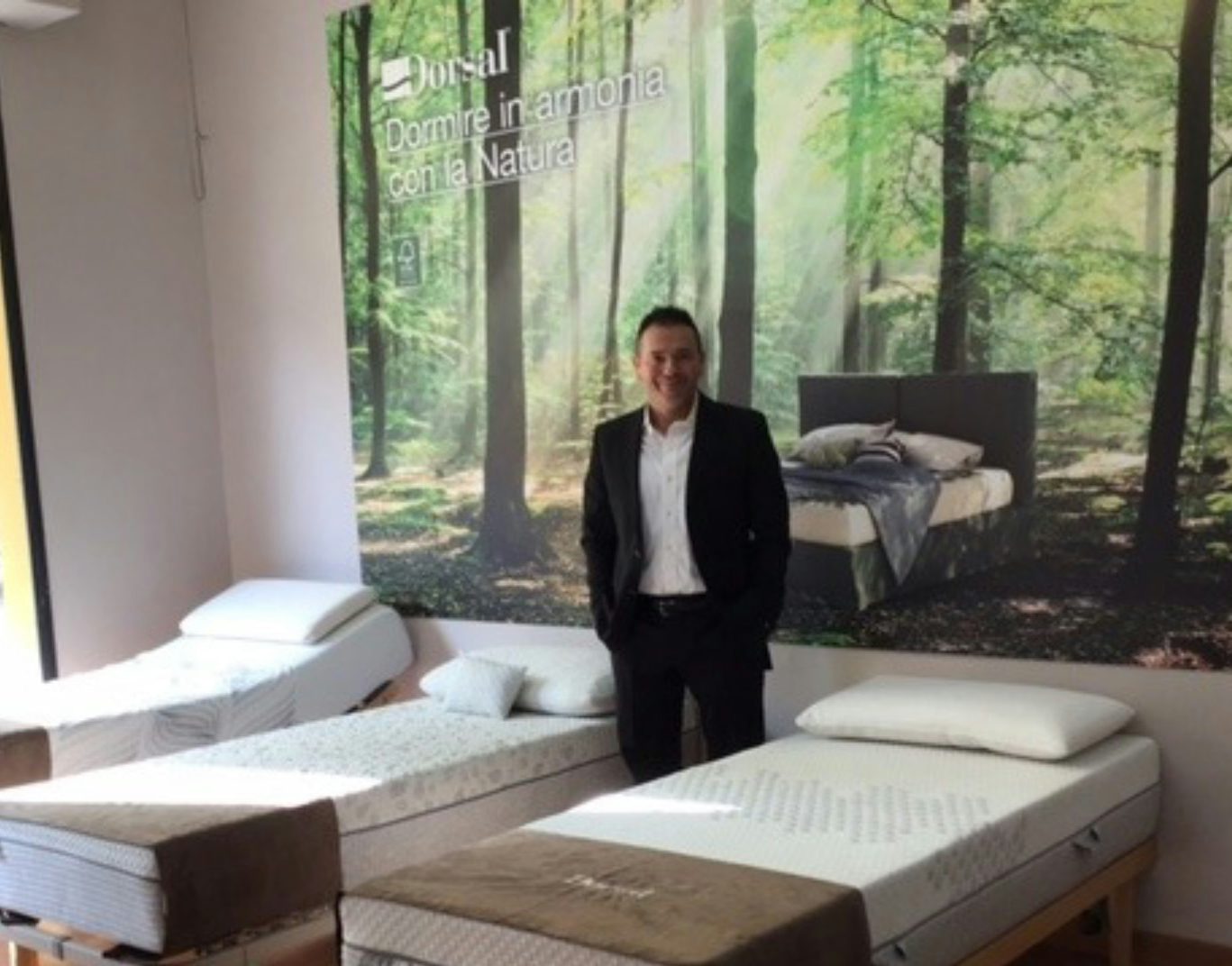 Uomo in completo nero in negozio di materassi con tre letti singoli e poster di foresta sullo sfondo.