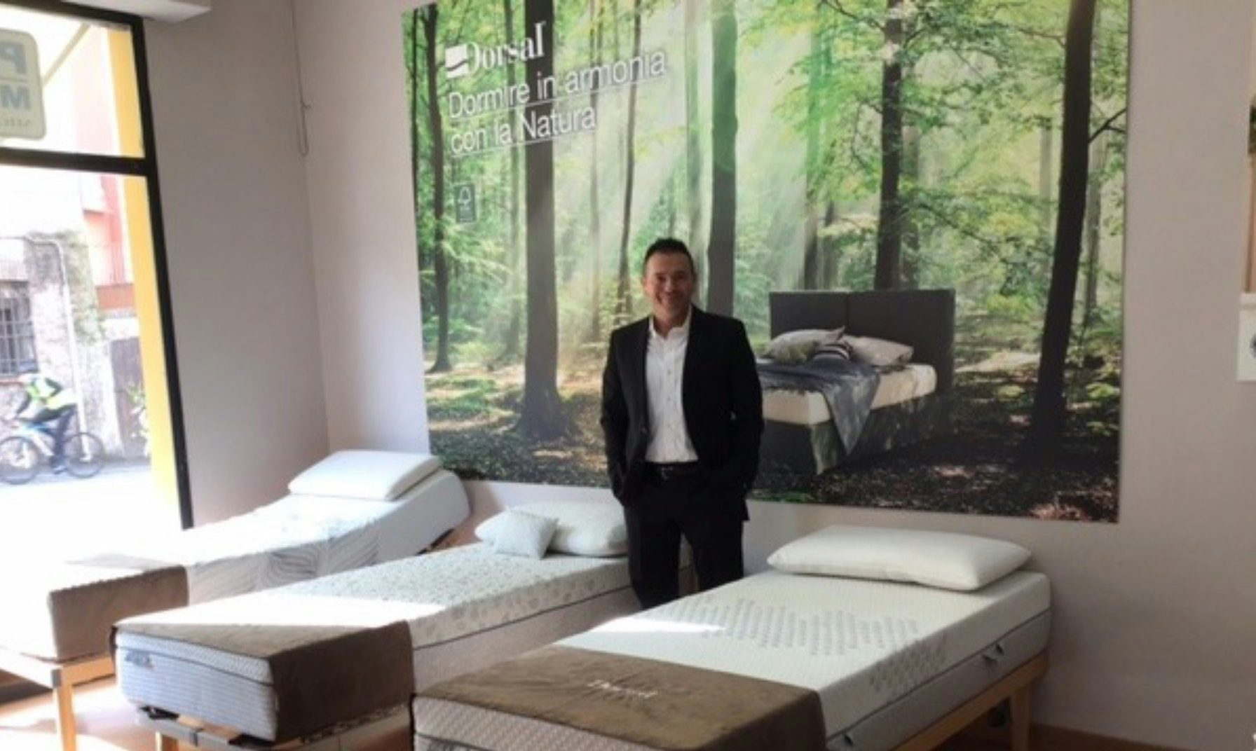 Uomo in completo nero in negozio di materassi con tre letti singoli e poster di foresta sullo sfondo.
