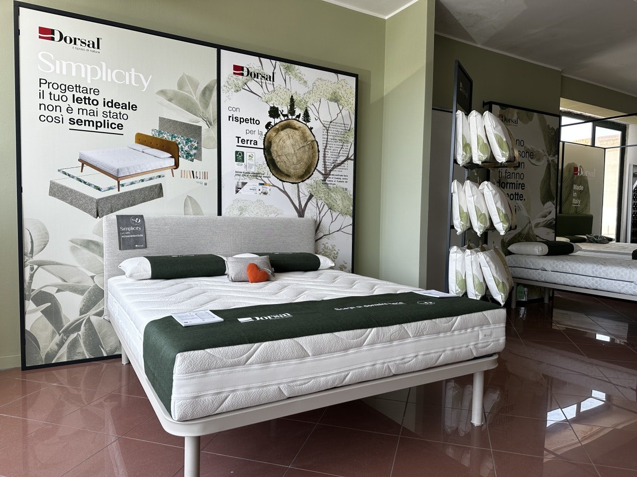 Letto matrimoniale con materasso Dorsal e cuscini bianchi in esposizione in negozio con parete verde chiaro e pavimento…