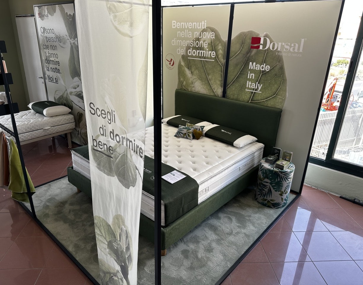 Letto matrimoniale con testiera verde, materasso bianco, cuscini e plaid verde, esposizione Dorsal con scritte promozionali.