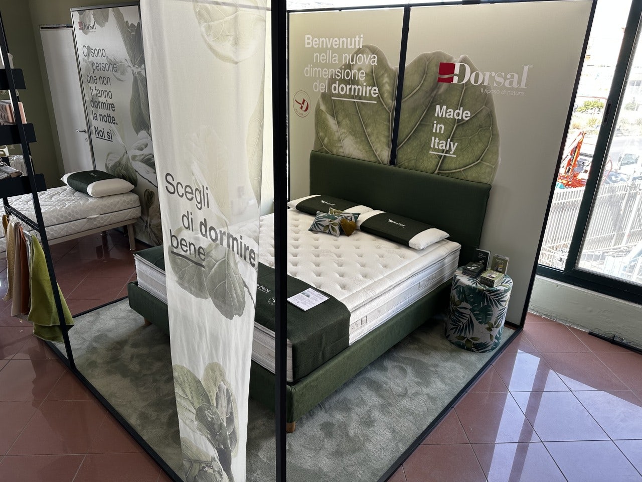 Letto matrimoniale con testiera verde, materasso bianco, cuscini e plaid verde, esposizione Dorsal con scritte promozionali.