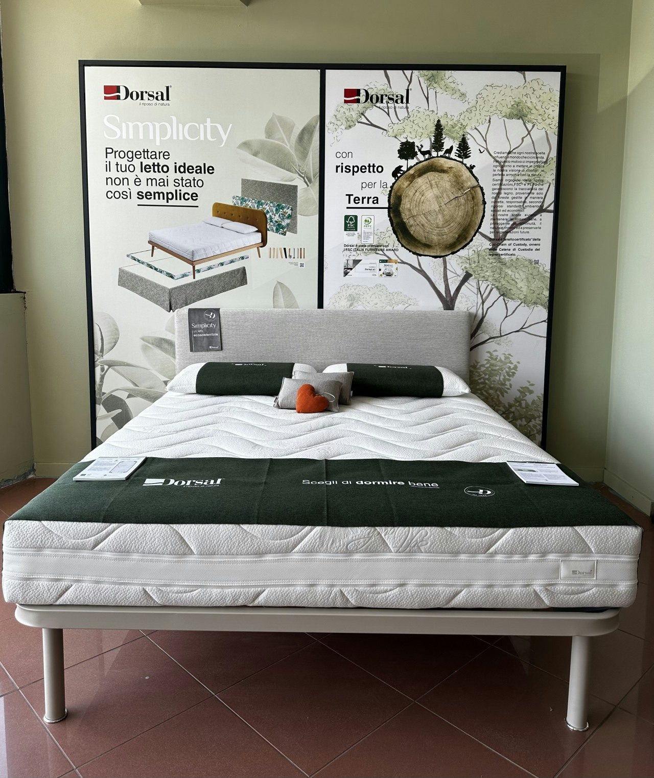Letto con testiera grigia, materasso bianco, cuscini verdi e due poster pubblicitari di Dorsal sullo sfondo.