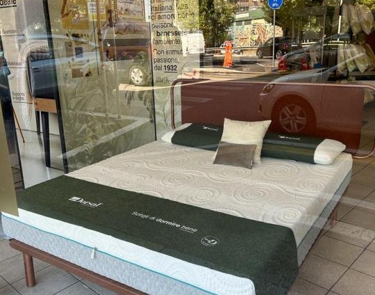 Materasso con copertura bianca e verde in esposizione su una struttura in legno dentro un negozio con riflessi della strada.