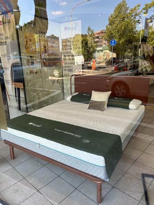 Materasso con copertura bianca e verde in esposizione su una struttura in legno dentro un negozio con riflessi della strada.