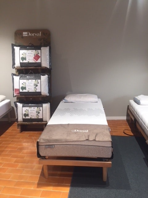 Letto singolo con materasso e cuscino Dorsal, scaffale con confezioni di cuscini e copriletto beige, pavimento in cotto.