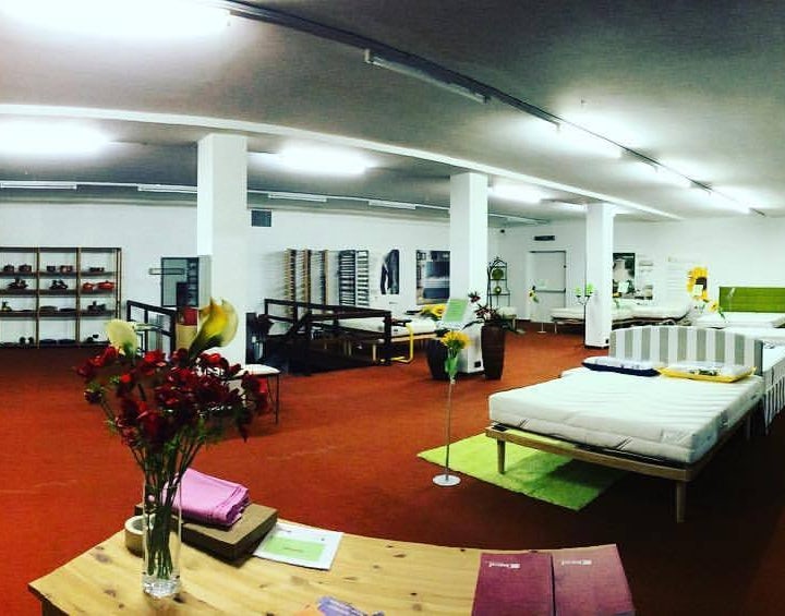 Negozio con letti esposti su moquette rossa, scaffali in legno, fiori e luci al neon sul soffitto bianco.