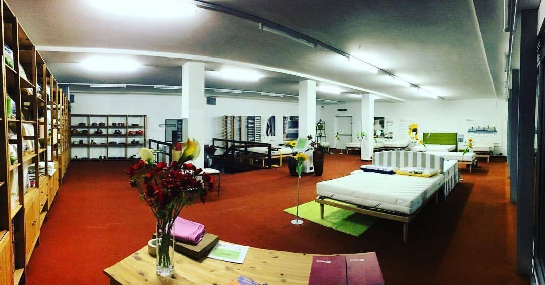 Negozio con letti esposti su moquette rossa, scaffali in legno, fiori e luci al neon sul soffitto bianco.