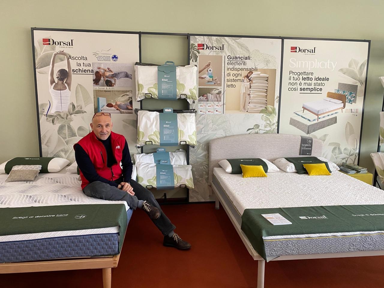 Uomo con giacca rossa seduto su letto in esposizione con cuscini e materassi Dorsal e pannelli informativi sul riposo.