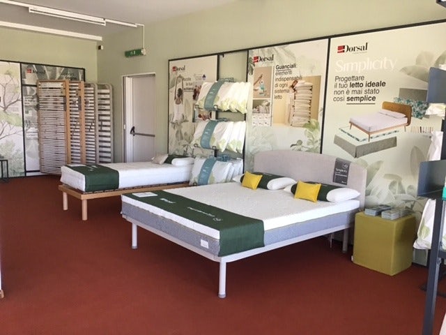Letti espositivi con materassi, cuscini e reti ortopediche in showroom con pavimento rosso e pareti chiare.