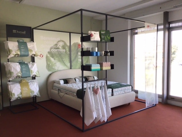 Letto imbottito con materasso, cuscini e coperte in esposizione in negozio con scaffali e accessori per il sonno.