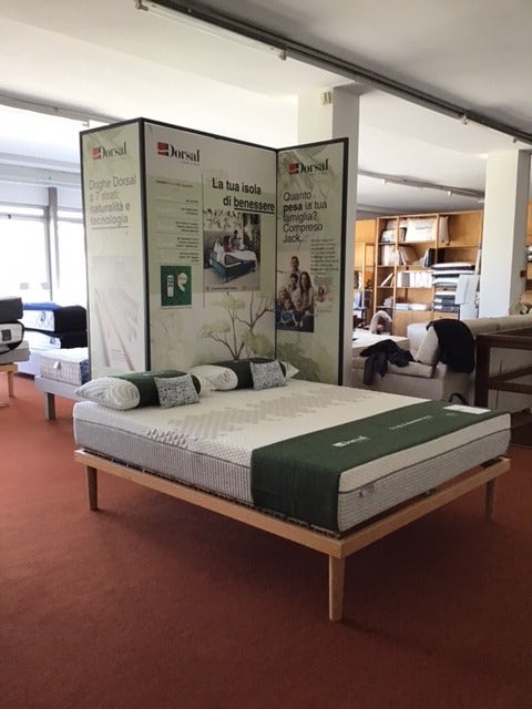 Letto matrimoniale con materasso Dorsal e cuscini, esposto su pavimento marrone in showroom con parete divisoria informativa.