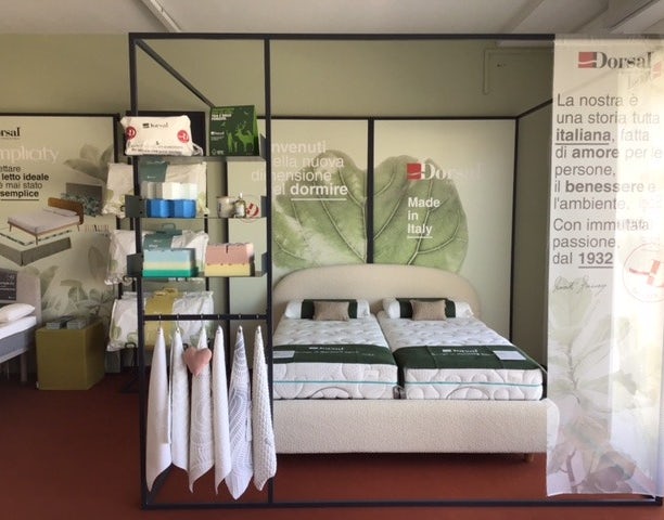 Letto matrimoniale con due materassi, scaffale con asciugamani e prodotti, sfondo con foglie e scritte in italiano.