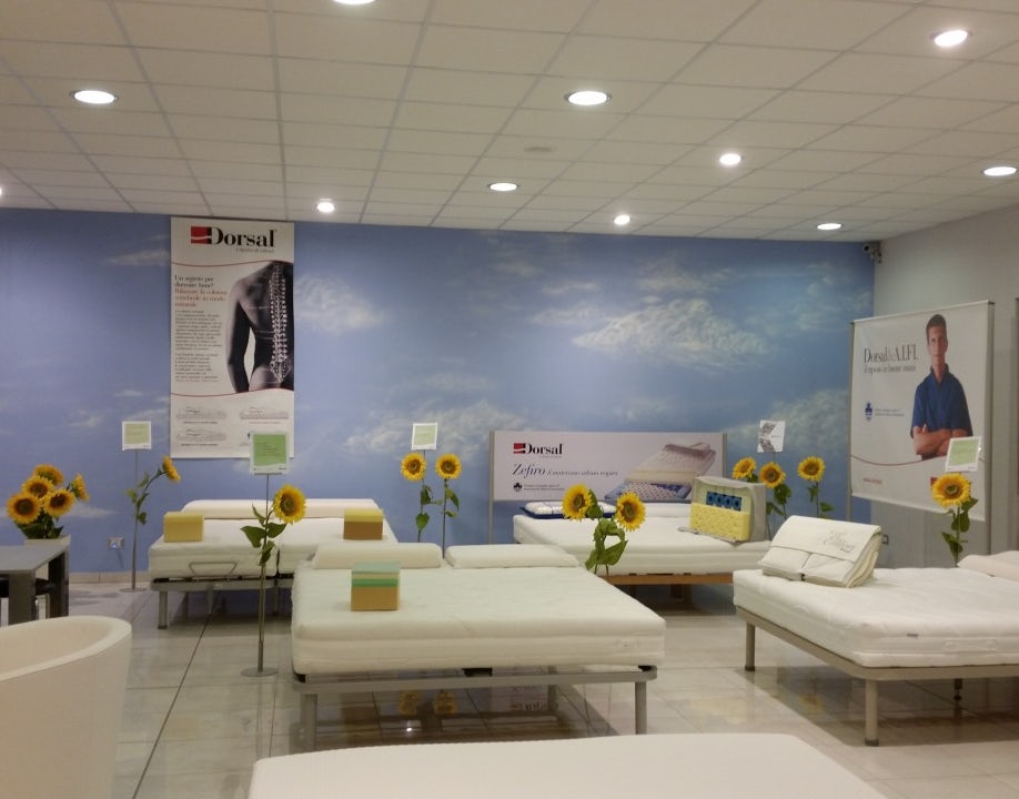 Negozio con letti ortopedici Dorsal, girasoli decorativi e donna seduta a scrivania, parete con cielo azzurro e nuvole.