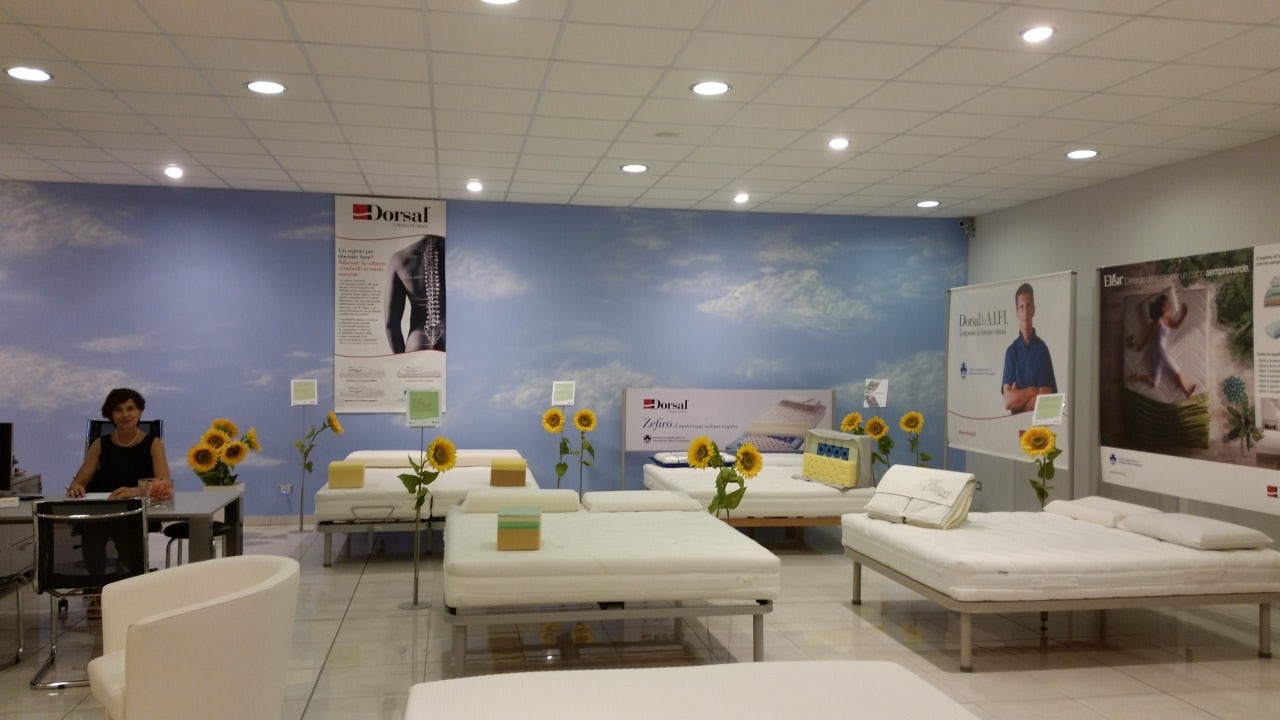 Negozio con letti ortopedici Dorsal, girasoli decorativi e donna seduta a scrivania, parete con cielo azzurro e nuvole.