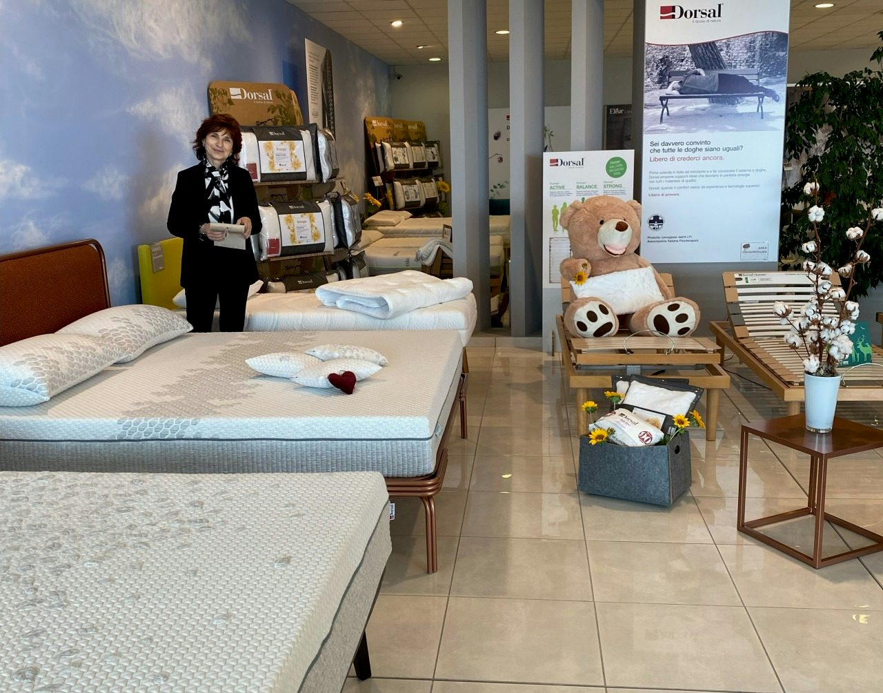 Negozio di materassi con donna sorridente, materassi esposti, orso di peluche su letto regolabile e insegne Dorsal.