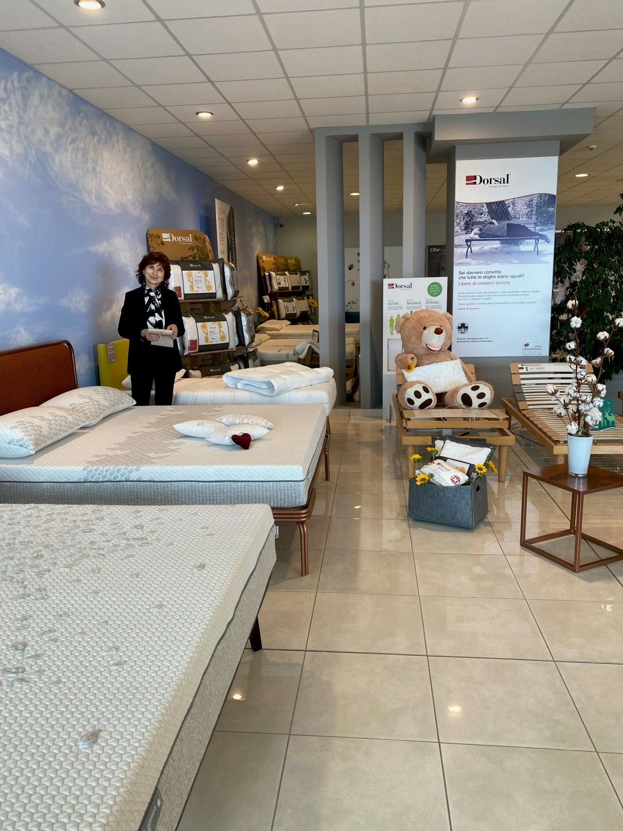 Negozio di materassi con donna sorridente, materassi esposti, orso di peluche su letto regolabile e insegne Dorsal.