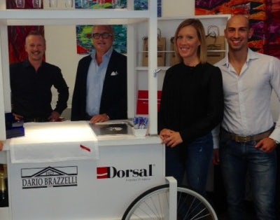 Quattro persone in piedi accanto a un carretto bianco con ruota anteriore, sopra scritto G.H. Mumm e Dario Brazzelli.