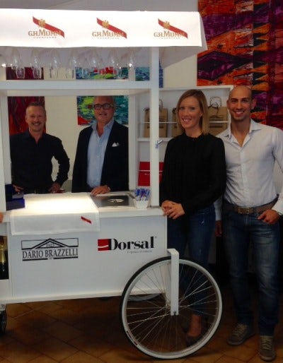 Quattro persone in piedi accanto a un carretto bianco con ruota anteriore, sopra scritto G.H. Mumm e Dario Brazzelli.