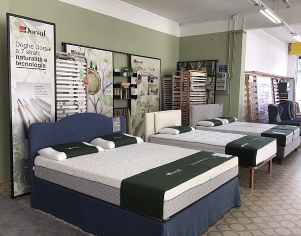 Esposizione di letti singoli e matrimoniali con materassi bianchi su strutture imbottite e cataloghi Dorsal sullo sfondo.