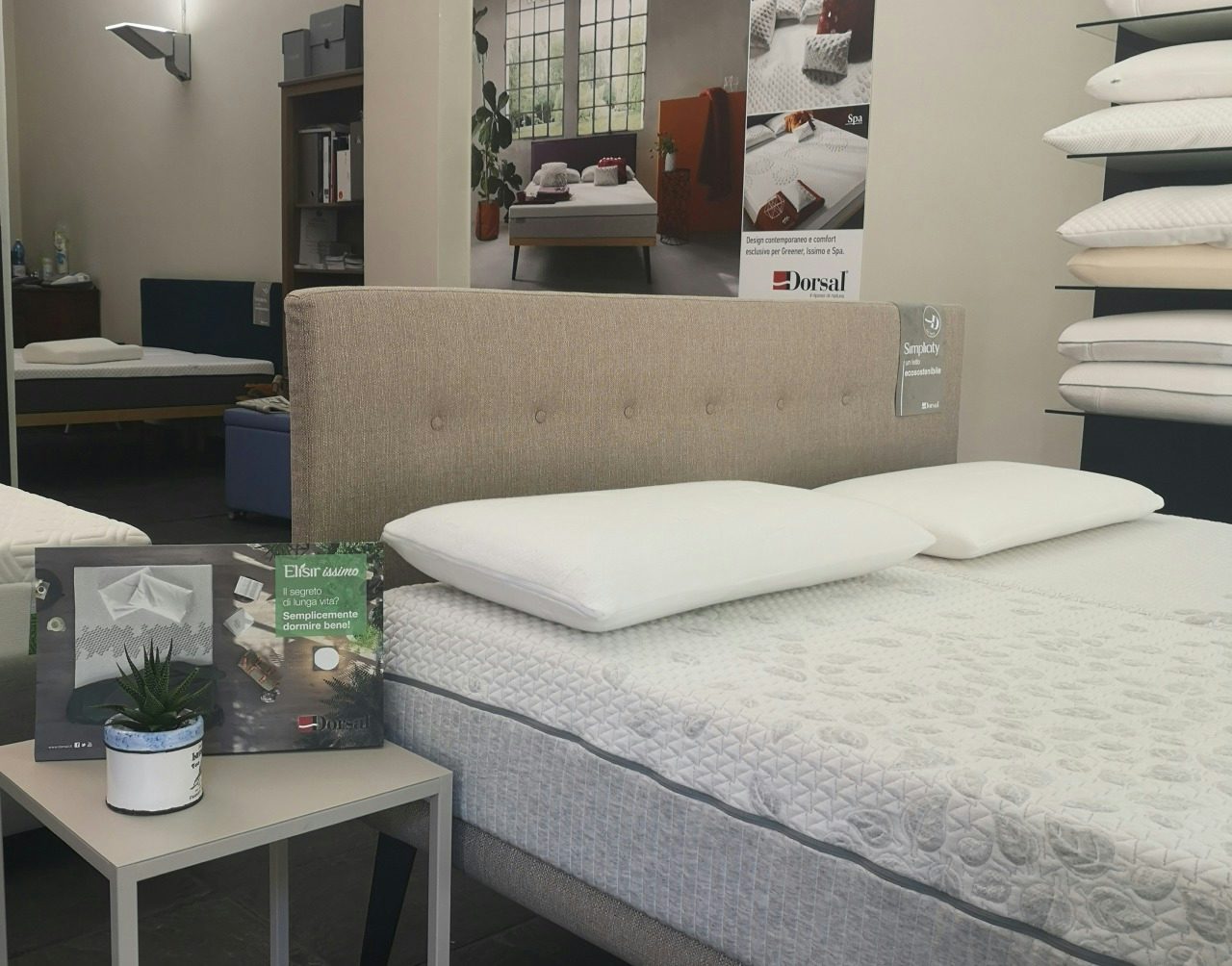 Letto matrimoniale beige con materasso e due cuscini bianchi, tavolino con pianta e brochure in negozio di arredamento per…