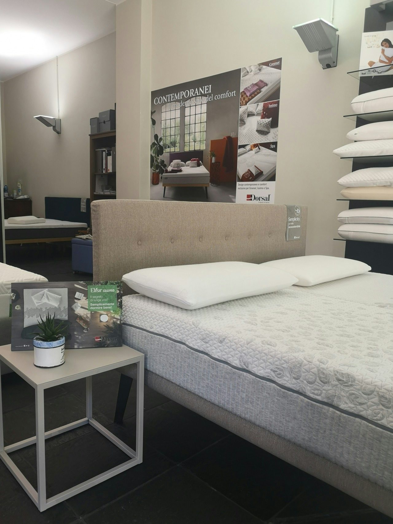 Letto matrimoniale beige con materasso e due cuscini bianchi, tavolino con pianta e brochure in negozio di arredamento per…