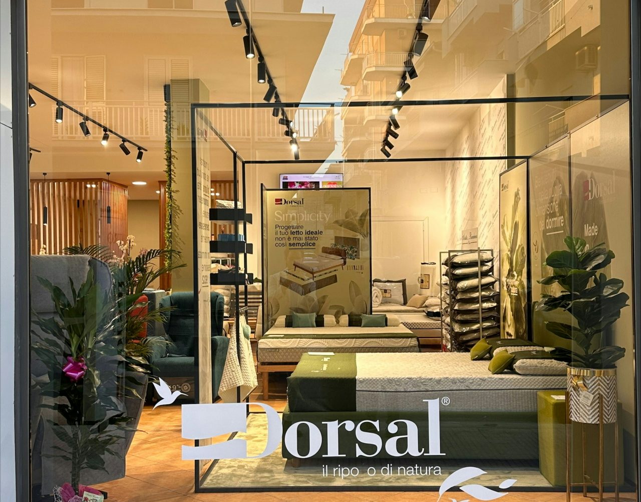 Vetrina di negozio con letti e materassi, piante decorative e poster con testo Dorsal il riposo di natura.