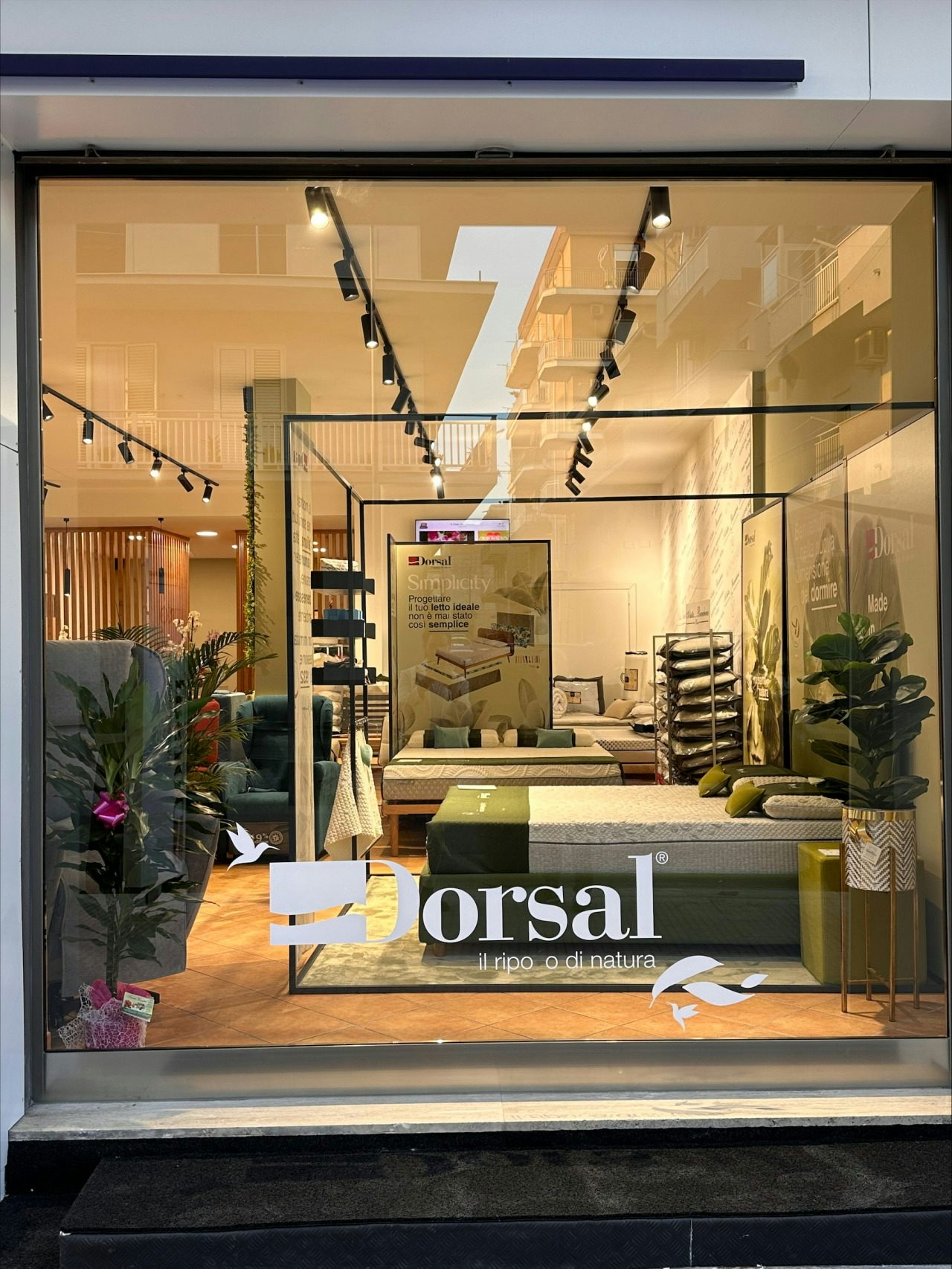 Vetrina di negozio con letti e materassi, piante decorative e poster con testo Dorsal il riposo di natura.