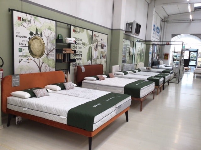 Agoni letti matrimoniali con testiere di diversi colori in esposizione in showroom con poster promozionali alle pareti.