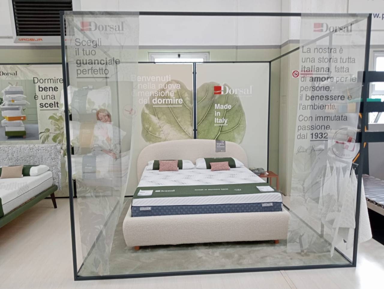 Letto matrimoniale con testiera curva imbottita beige in esposizione, sfondo con grandi foglie e scritte promozionali in…