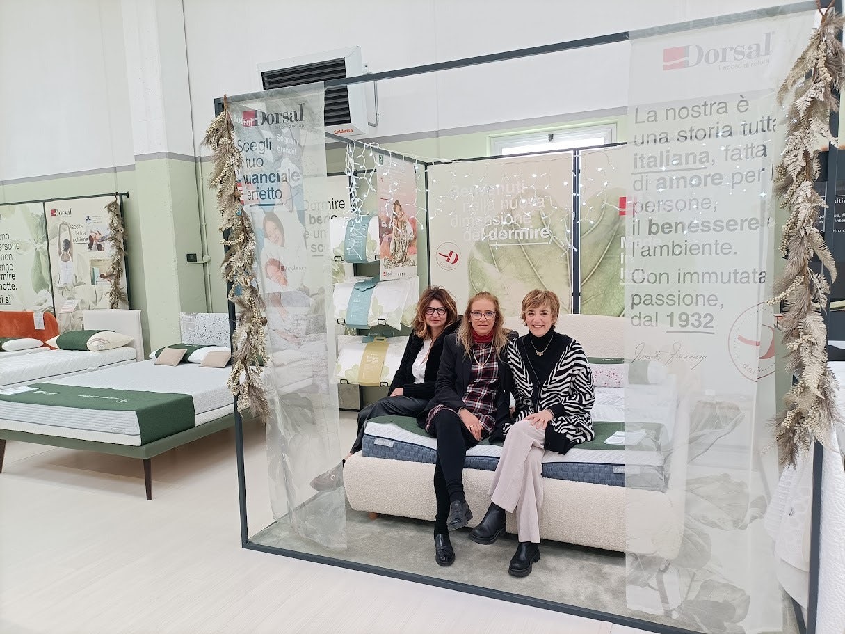 Tre donne sedute su un letto in esposizione in un negozio di materassi Dorsal con decorazioni di felci e testo promozionale.