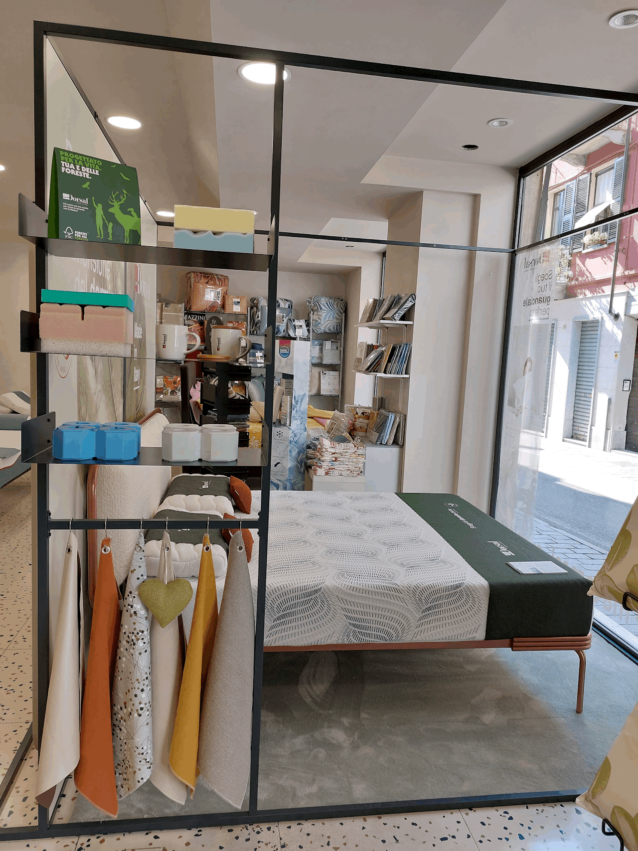 Letto matrimoniale con materasso fogliato grigio e rete in metallo, accanto scaffale espositivo con tessuti colorati appesi.