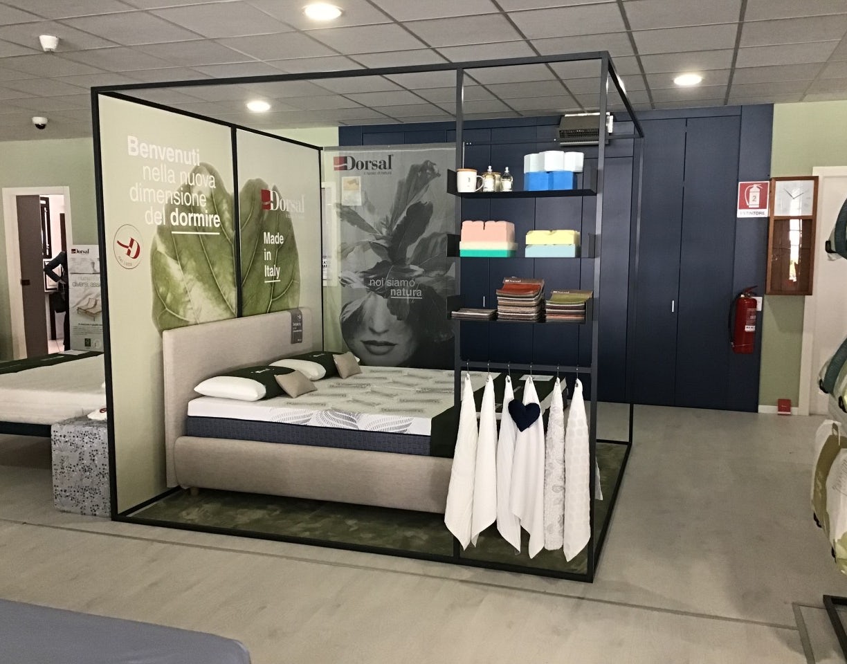 Letto matrimoniale in esposizione con cuscini, tessuti appesi e cartelloni pubblicitari con scritte Benvenuti e made in…