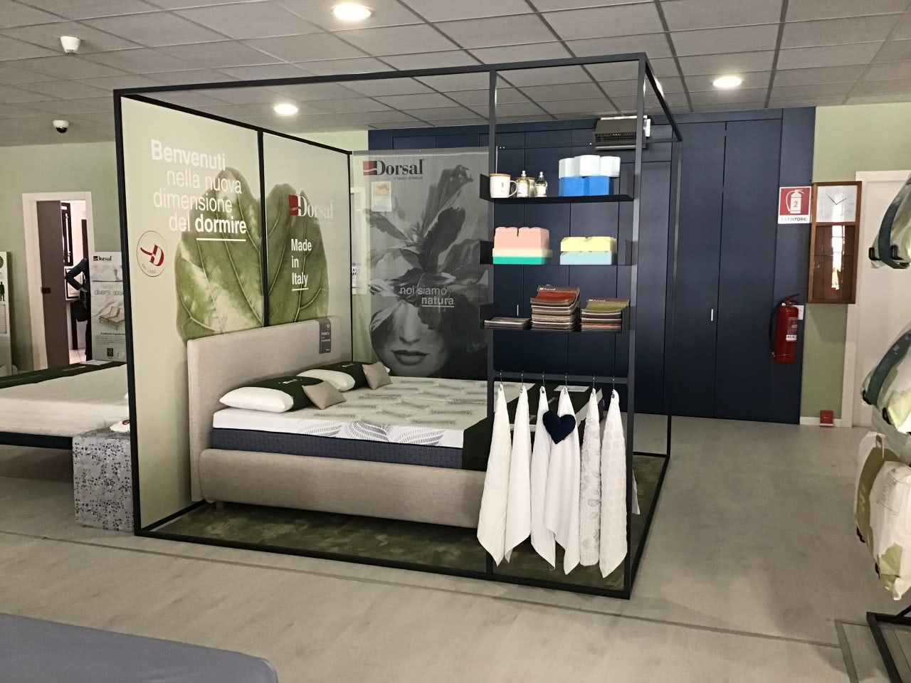 Letto matrimoniale in esposizione con cuscini, tessuti appesi e cartelloni pubblicitari con scritte Benvenuti e made in…