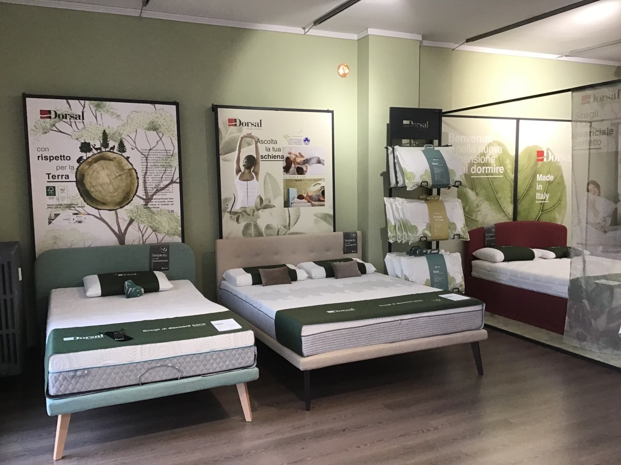 Tre letti con materassi Dorsal in esposizione con poster pubblicitari e cuscini coordinati in una stanza con pavimento in…